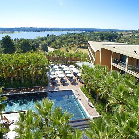 Ap Lago Montargil - Conference & Spa Exterior foto The photo shows a scenic view of a modern hotel or resort. In the foreground, there is a swimming pool surrounded by palm trees, with lounge chairs positioned beside it. Sun umbrellas provide shade over the chairs. The background features a lush land