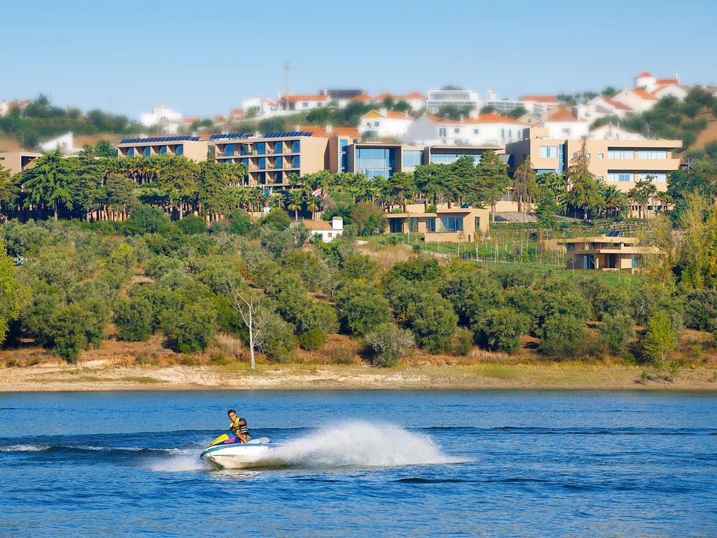 Ap Lago Montargil - Conference & Spa Exterior foto The photo depicts a person riding a jet ski on a body of water. In the background, there are modern buildings and lush greenery, suggesting a scenic landscape, likely near a resort or upscale residential area. The clear blue sky and calm water enhanc