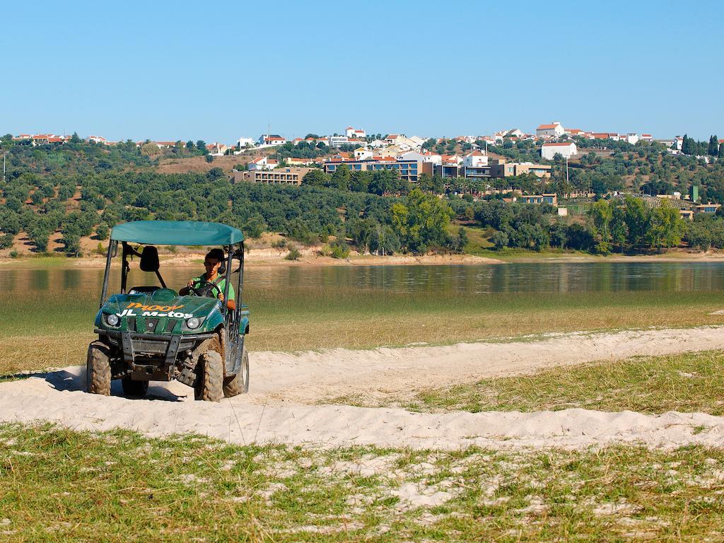 Ap Lago Montargil - Conference & Spa Exterior foto The photo shows a small green all-terrain vehicle driving along a sandy path beside a body of water. In the background, there's a landscape with trees and rolling hills, along with a town or village visible on the hillside. The sky is clear, indicati