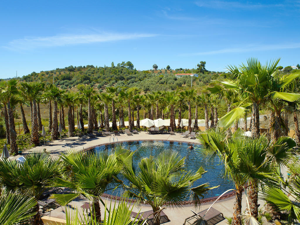 Ap Lago Montargil - Conference & Spa Exterior foto The photo depicts a beautiful outdoor area featuring a circular swimming pool surrounded by palm trees. The pool area has several lounge chairs arranged around it. In the background, there is a lush green hillside, and the sky is clear with a bright 