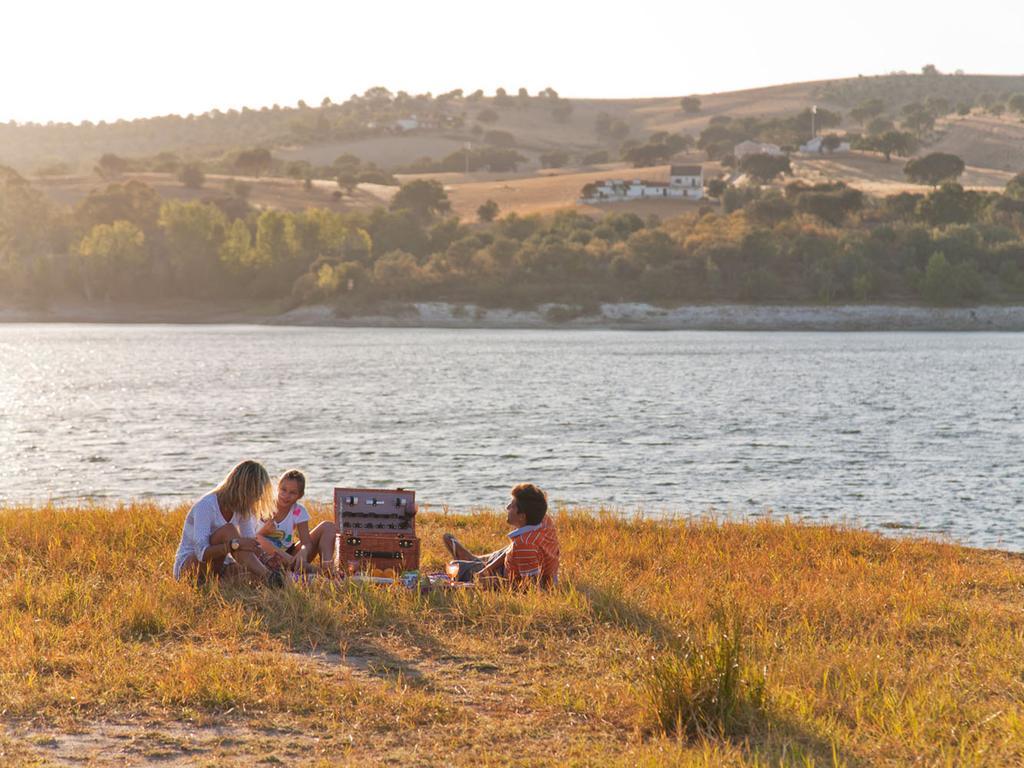 Ap Lago Montargil - Conference & Spa Exterior foto The photo depicts a serene lakeside scene during sunset. Three people are gathered on a grassy area near the water, enjoying a picnic. They are seated around a picnic basket, with the shimmering lake in the background and hills in the distance. The w