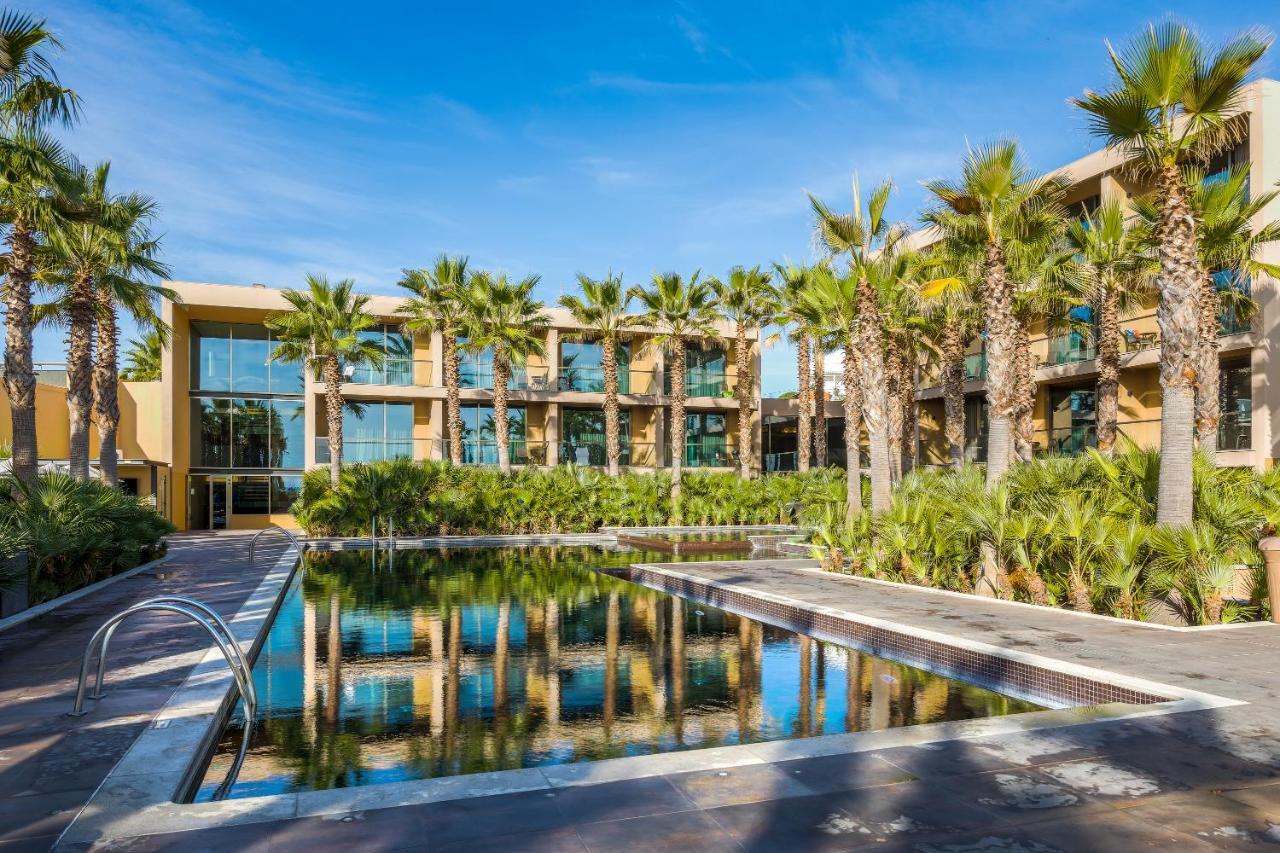 Ap Lago Montargil - Conference & Spa Exterior foto The photo features a modern building surrounded by lush greenery and palm trees. In the foreground, there is a serene swimming pool with a reflective surface that captures the surrounding environment. The architecture of the building appears to have 