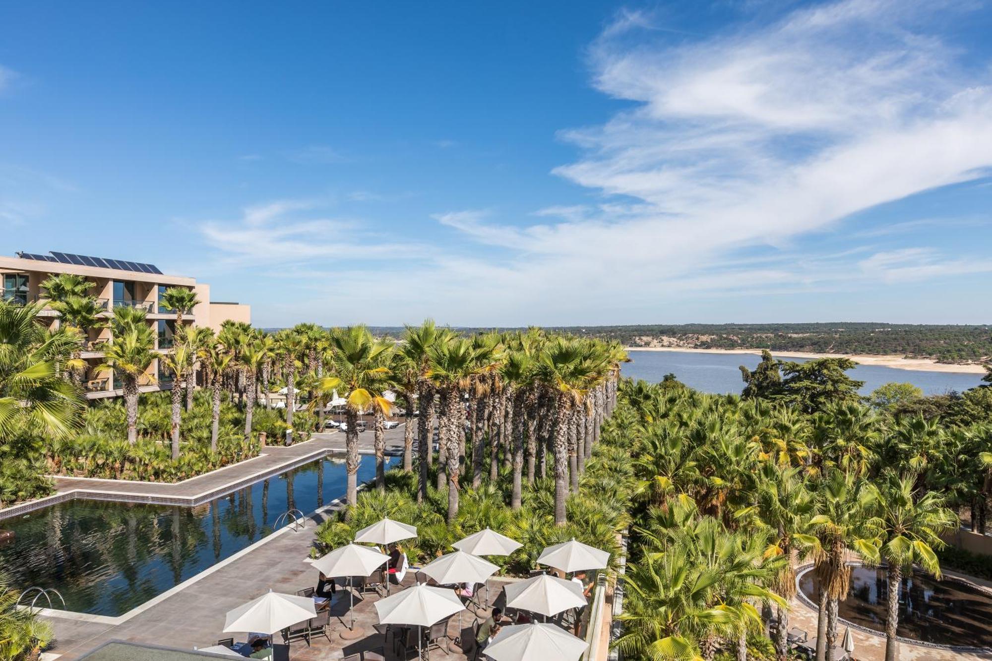Ap Lago Montargil - Conference & Spa Exterior foto The photo shows a scenic view of a resort or hotel area. In the foreground, there is a pool surrounded by palm trees and lounge chairs with umbrellas, suggesting a relaxing atmosphere. Beyond the pool, there is a body of water, likely a river or coas