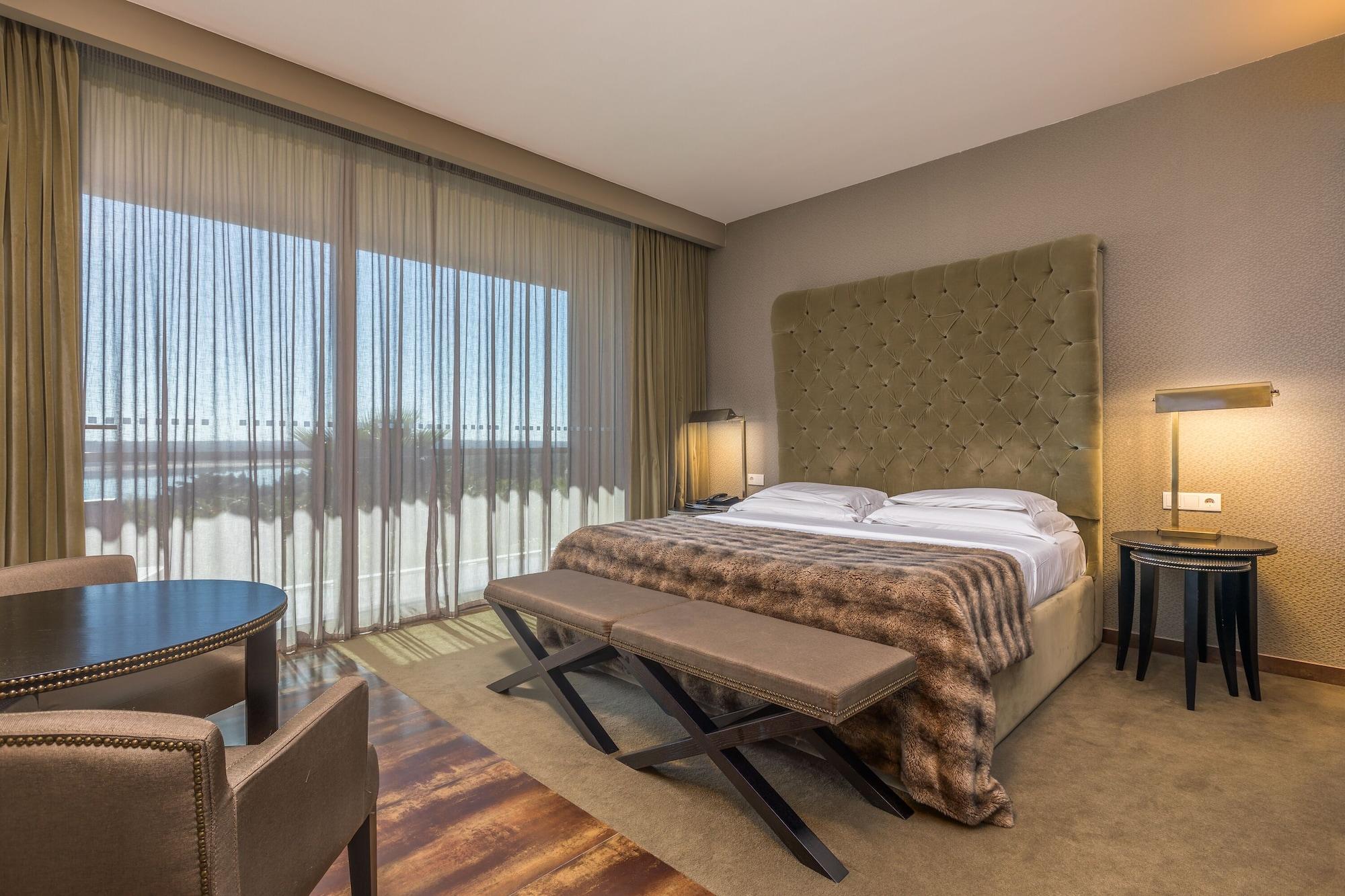 Ap Lago Montargil - Conference & Spa Exterior foto The photo shows a modern hotel room featuring a large bed with a tufted headboard and a cozy blanket. There are curtains draped over large windows that allow natural light to flow in, providing a view of the outside. A small seating area includes two