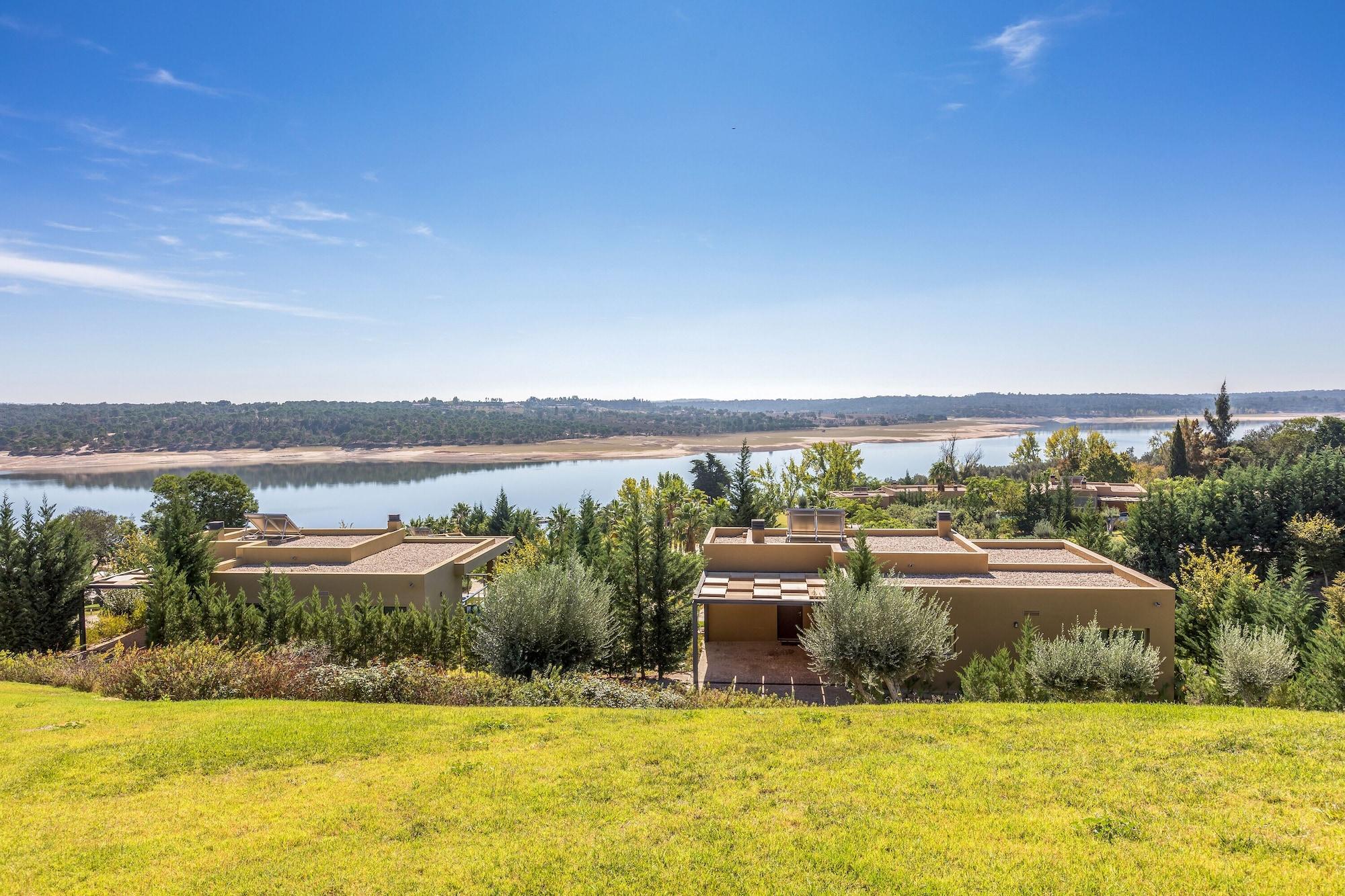 Ap Lago Montargil - Conference & Spa Exterior foto The photo depicts a scenic landscape featuring a calm body of water, possibly a lake or river, surrounded by lush greenery and trees. In the foreground, there are several modern buildings with flat roofs, set against a vibrant blue sky. The overall s