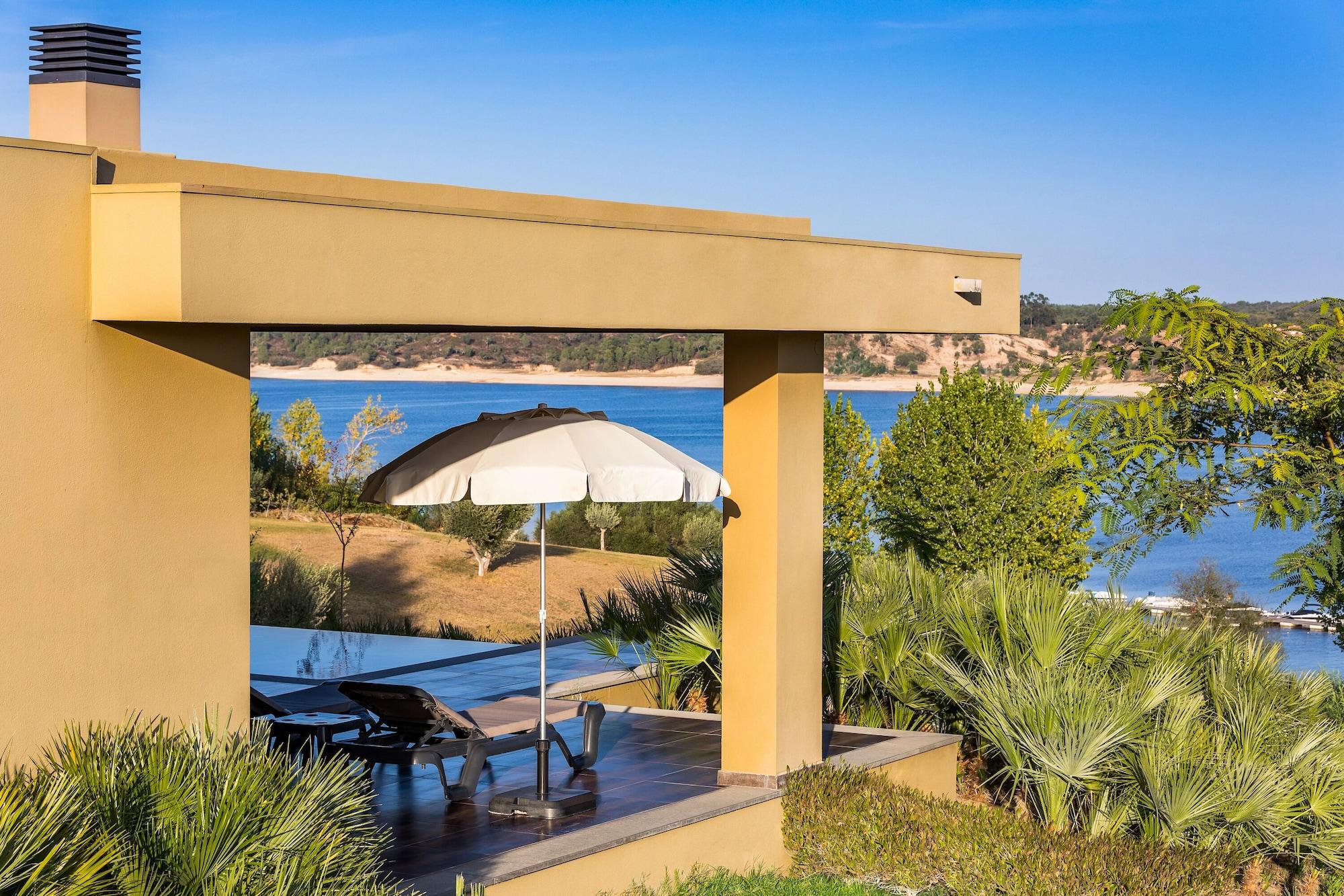 Ap Lago Montargil - Conference & Spa Exterior foto The photo shows a peaceful outdoor area with a modern architectural structure featuring an overhang. There is a lounge chair positioned beneath a large umbrella, creating a shaded spot to relax. In the background, there is a picturesque view of a bod