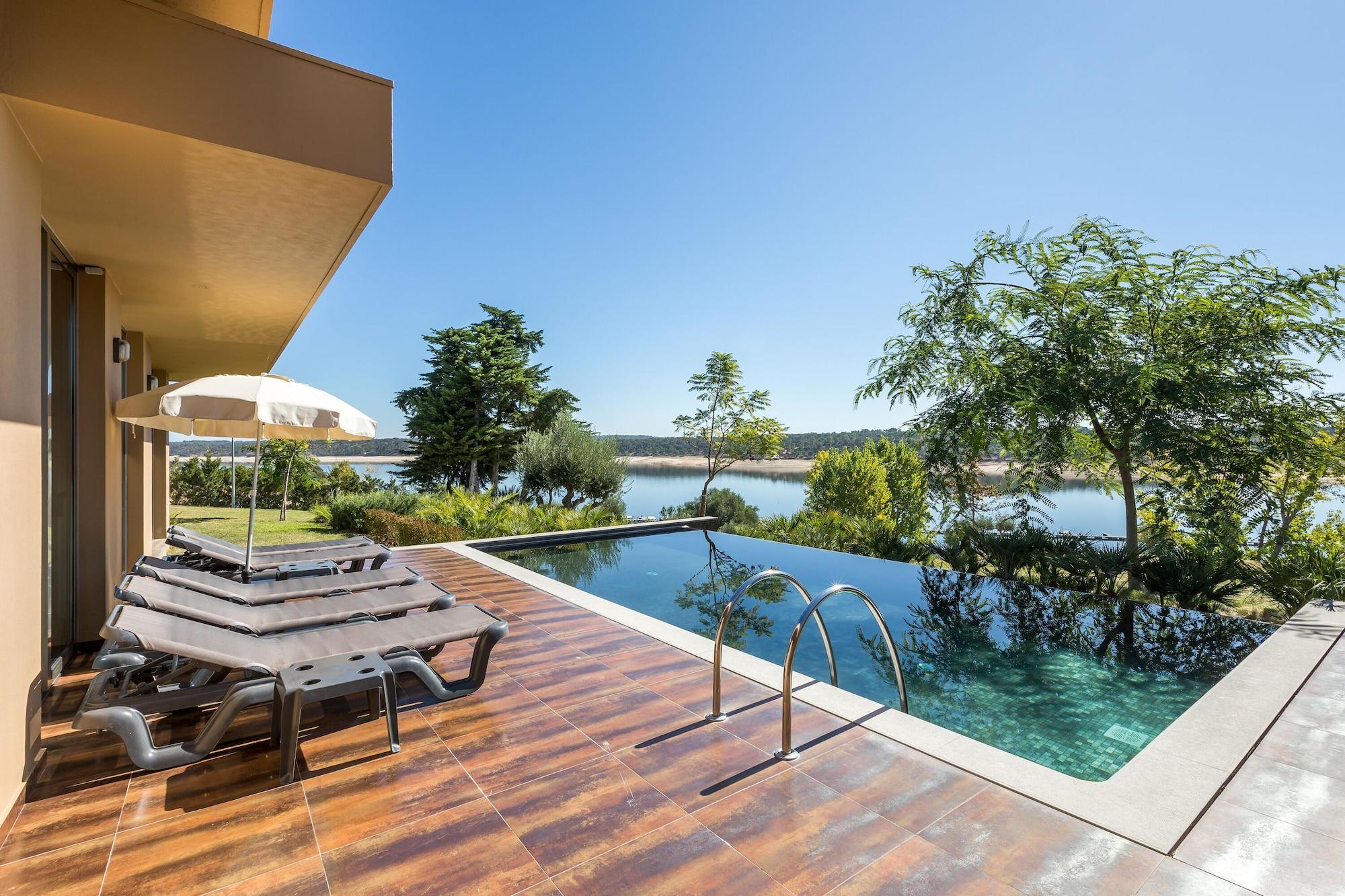 Ap Lago Montargil - Conference & Spa Exterior foto The photo shows a luxurious outdoor setting by a pool. There are several lounge chairs arranged next to an infinity pool, which overlooks a scenic body of water and lush greenery. A parasol provides shade for relaxation. The sky is clear and blue, in