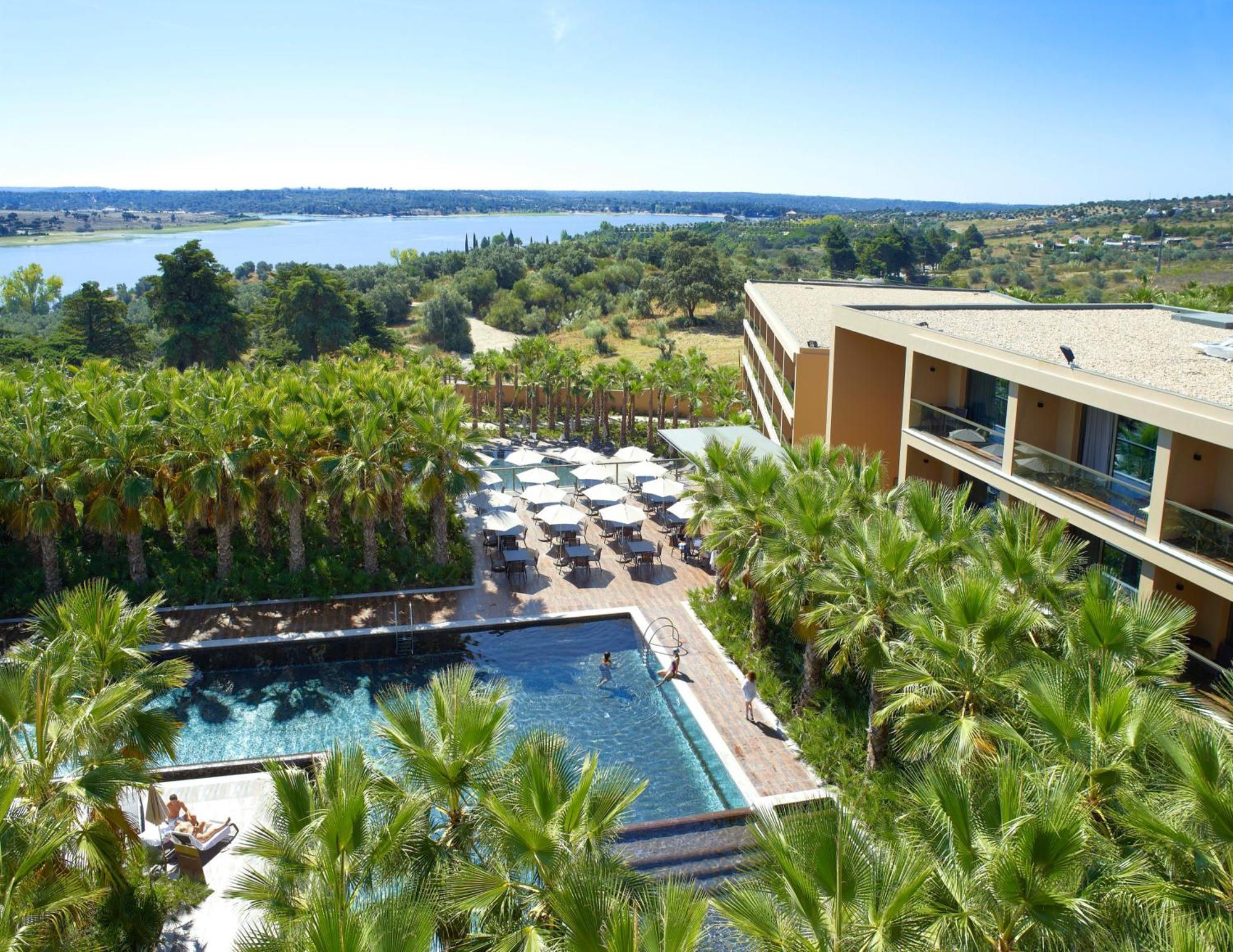 Ap Lago Montargil - Conference & Spa Exterior foto The photo shows a scenic view of a modern hotel or resort. In the foreground, there is a swimming pool surrounded by palm trees, with lounge chairs positioned beside it. Sun umbrellas provide shade over the chairs. The background features a lush land