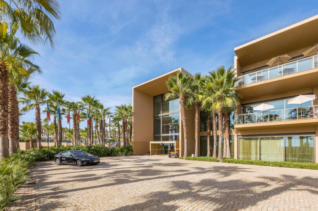 Ap Lago Montargil - Conference & Spa Exterior foto The photo shows a modern building with a sleek design, featuring large glass windows and balconies. The structure is surrounded by a well-maintained outdoor area with palm trees, adding a tropical feel. In front of the building, there is a paved park